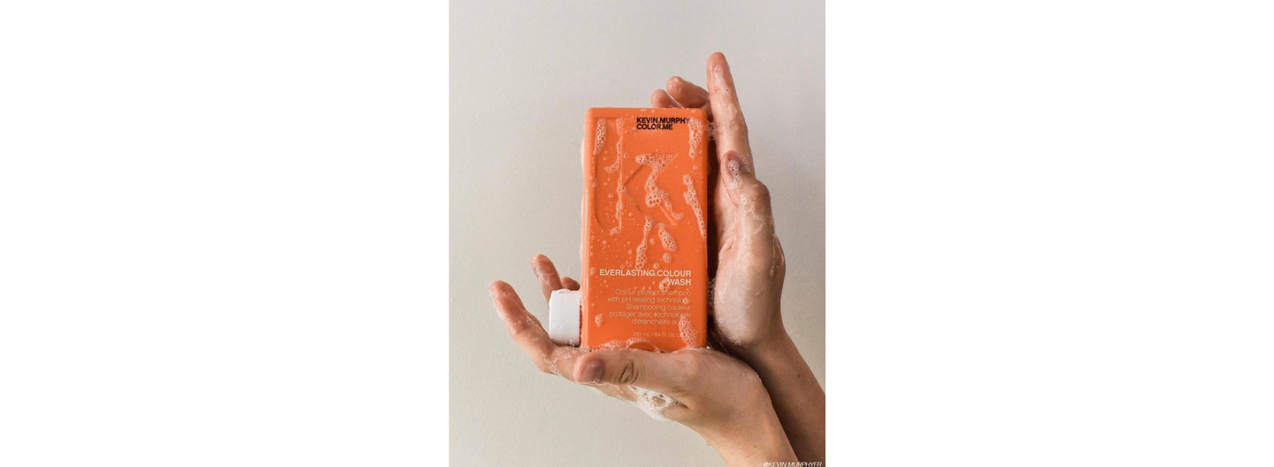 Hands holding a bottle of Kevin Murphy Everlasting Colour Wash shampoo against a neutral background, showcasing the product's details and foamy texture